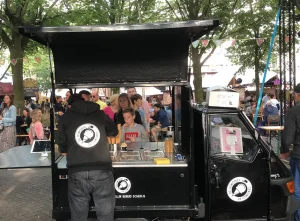 Ijsschepper hard aan het werk bij zijn ijskar van De IJsmobiel op een buitenfeest met een rij klanten wachtend aan de andere kant van de kar.